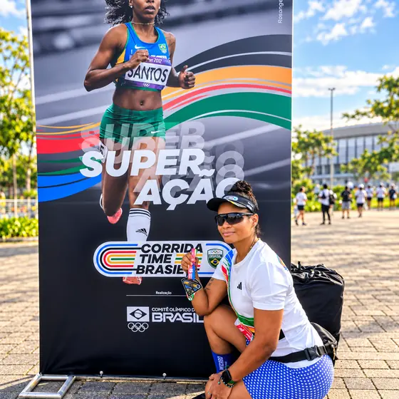Patrícia agachada com a medalha em frente ao painel Superação da Corrida Time Brasil